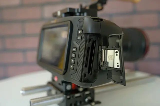 Blackmagic Pocket 6K Pro montée dans sa cage sur rails, trappe des cartes mémoire ouverte, devant un mur de briques.